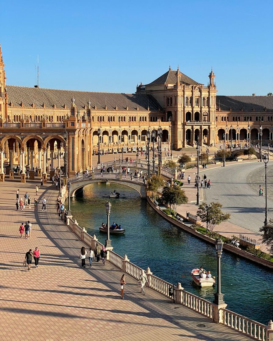 Canal semicircular de la plaza de España de Sevilla con una barca de remos y el puente de cerámica en primer plano, la torre norte al fondo bajo un cielo despejado